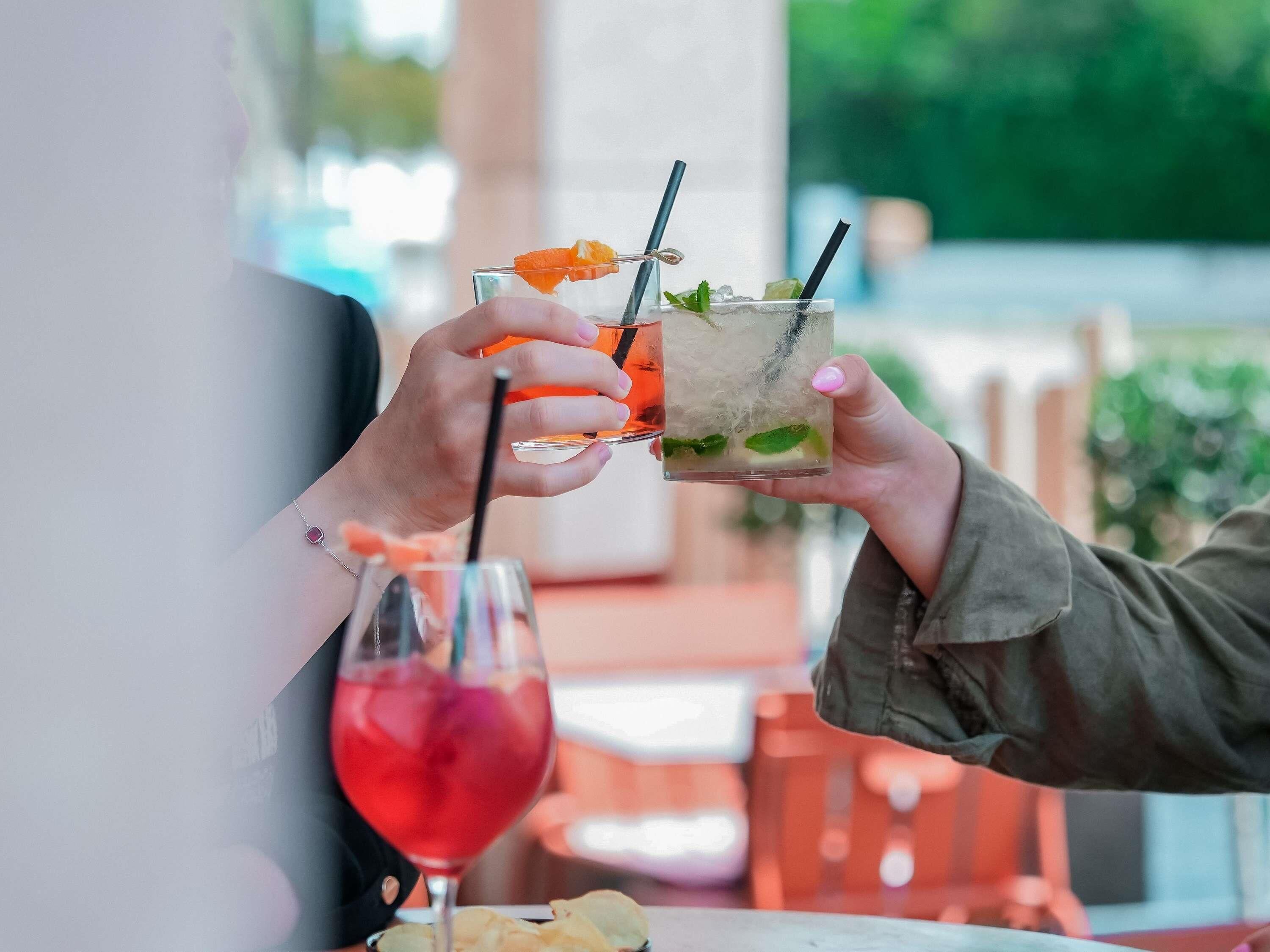 Dos personas sosteniendo vasos con bebidas, con una bebida roja al fondo.