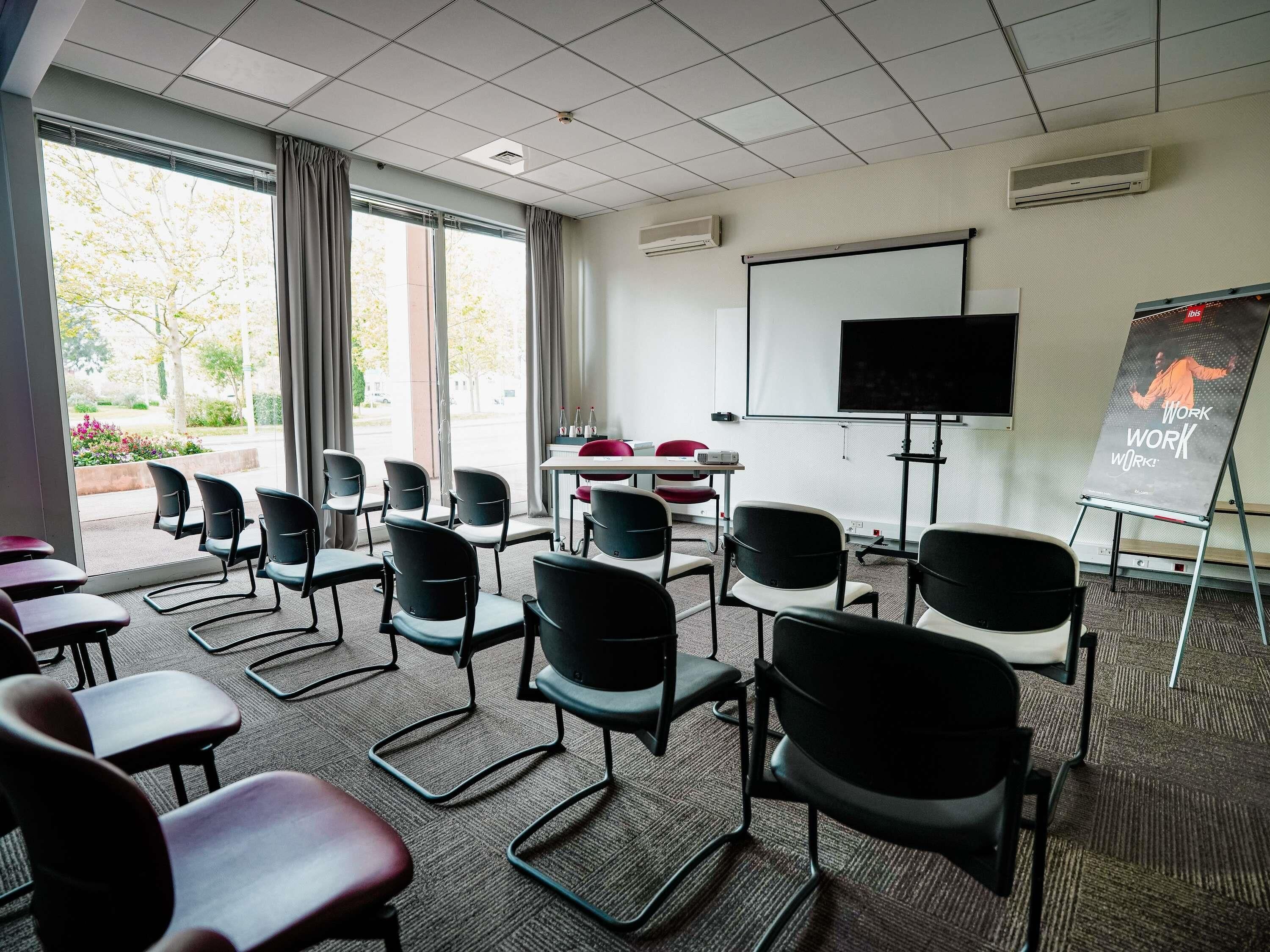 Une salle de conférence équipée d’un grand écran, d’un tableau blanc et de nombreuses chaises disposées autour de tables.