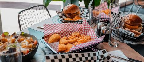 A table set with food items including burgers, fries, and onion rings, with checkered paper baskets and glasses.