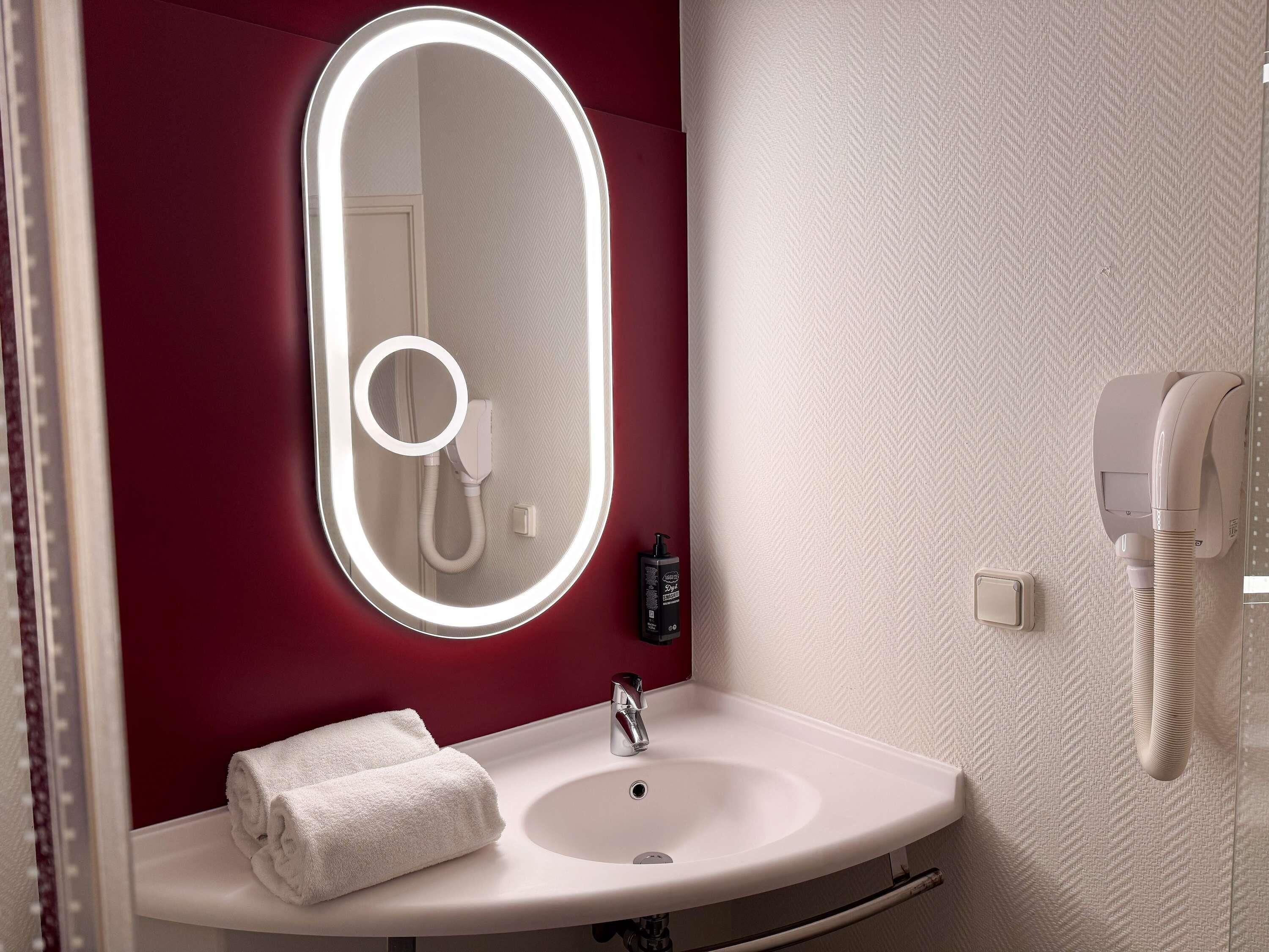 A bathroom with a white sink, a mirror with a light, a towel, and a hairdryer on the wall.