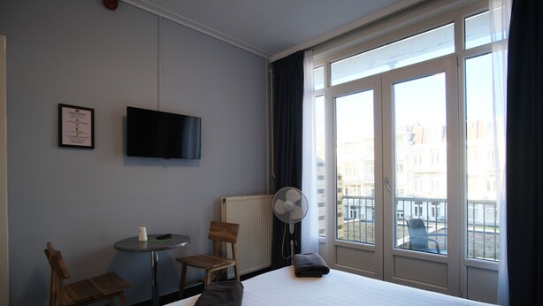 Habitación de hotel con cama, una silla, una mesita, televisor, ventana y ventilador, con vistas a edificios.
