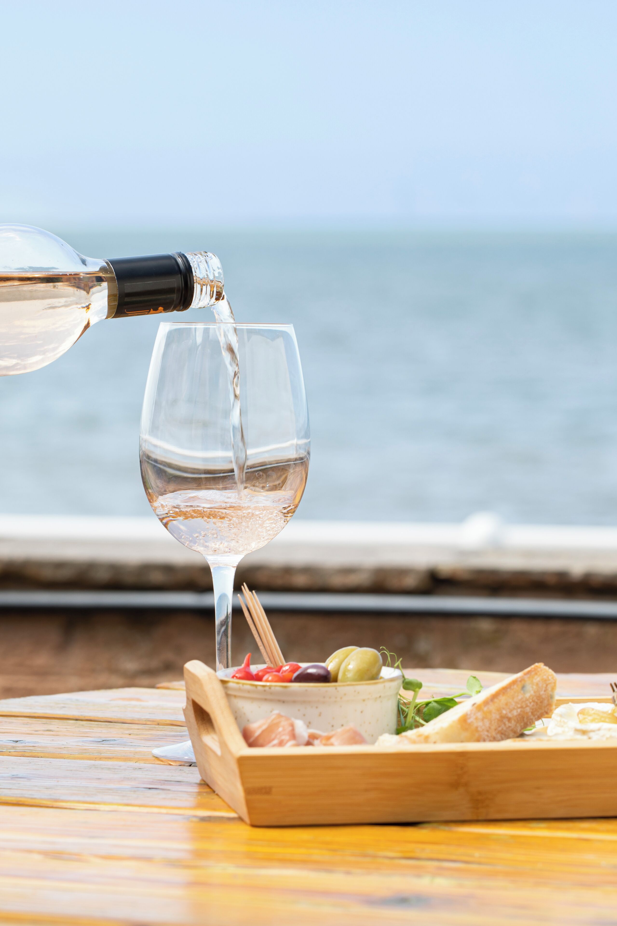 A glass of white wine being poured from a bottle, with a wooden board and food items in the foreground, and a view of the ocean in the background.