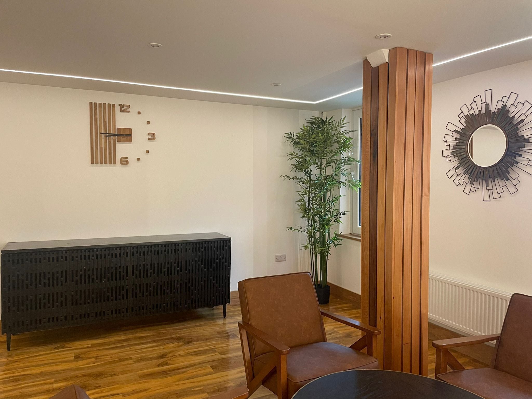 Een moderne kamer met een zwarte console tafel, twee bruine stoelen, een hoge bamboe plant en een decoratieve spiegel aan de muur.