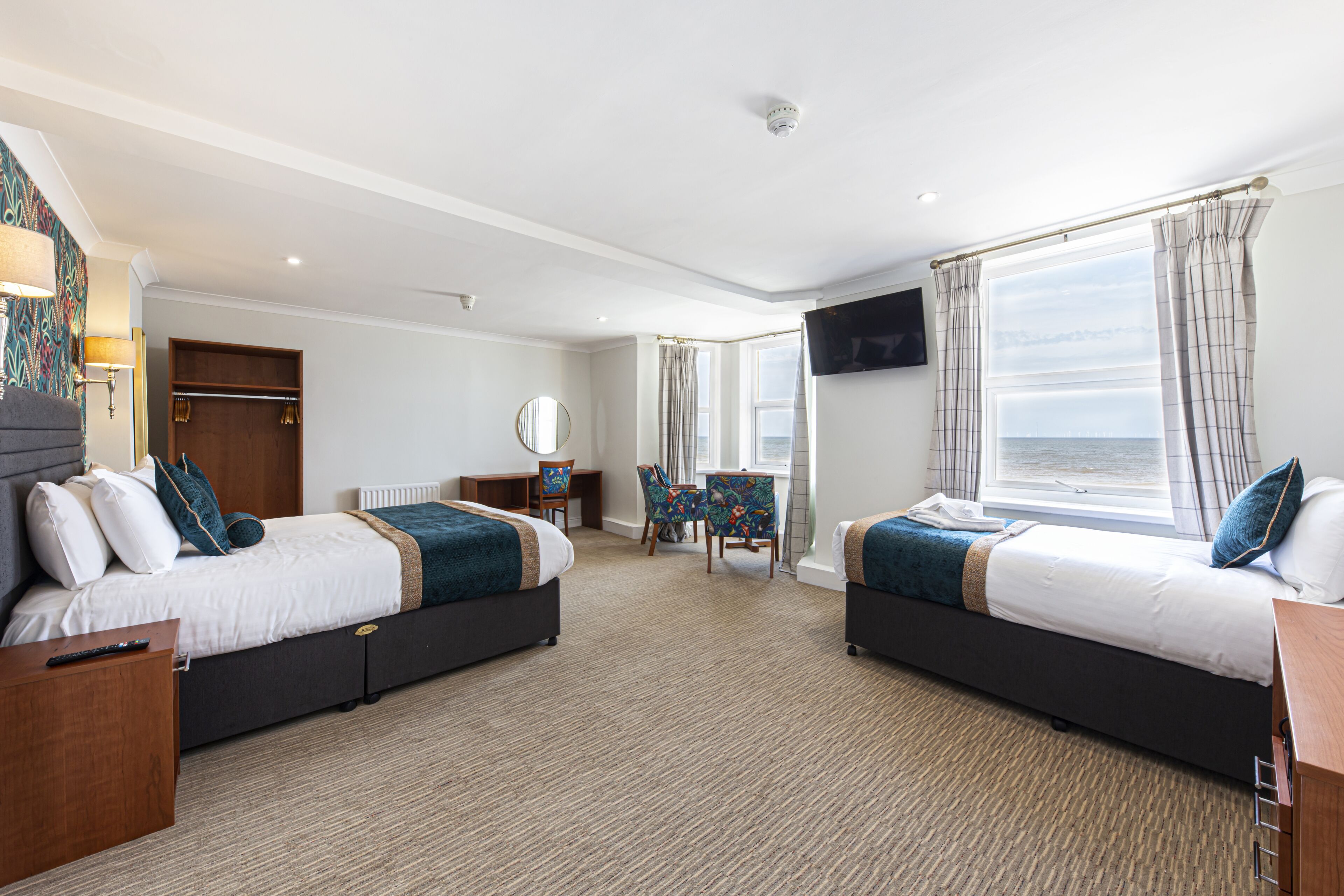 A hotel room with two beds, a TV, a desk, a chair, and a view of the sea.