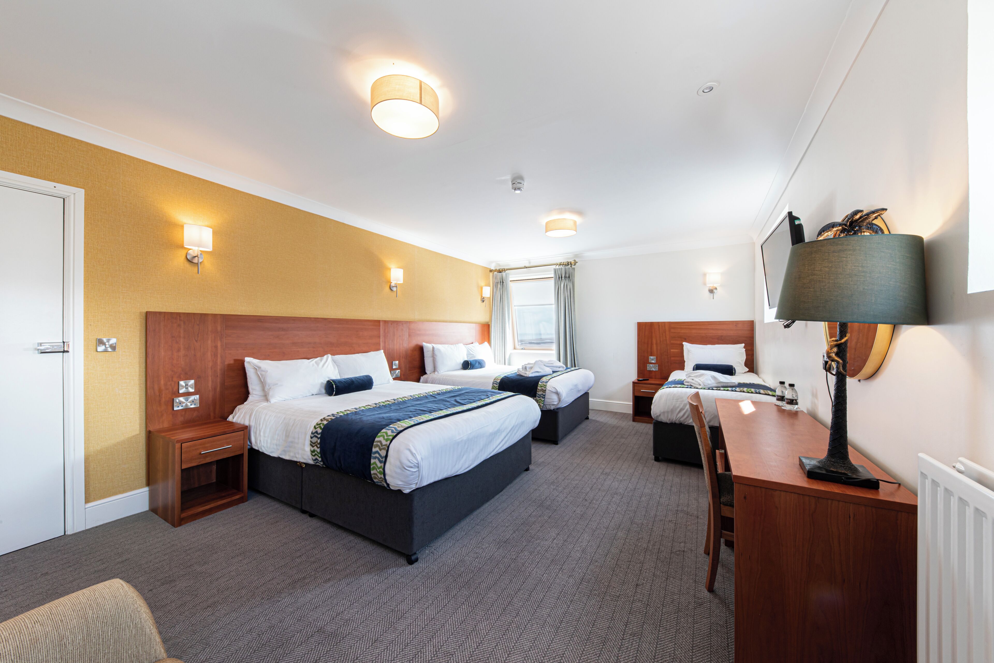 Quarto de hotel com duas camas, uma escrivaninha, uma cadeira, um abajur e uma janela com cortinas.