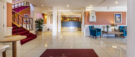 A hotel lobby with a red carpet, a staircase, a reception desk, and a bar area.
