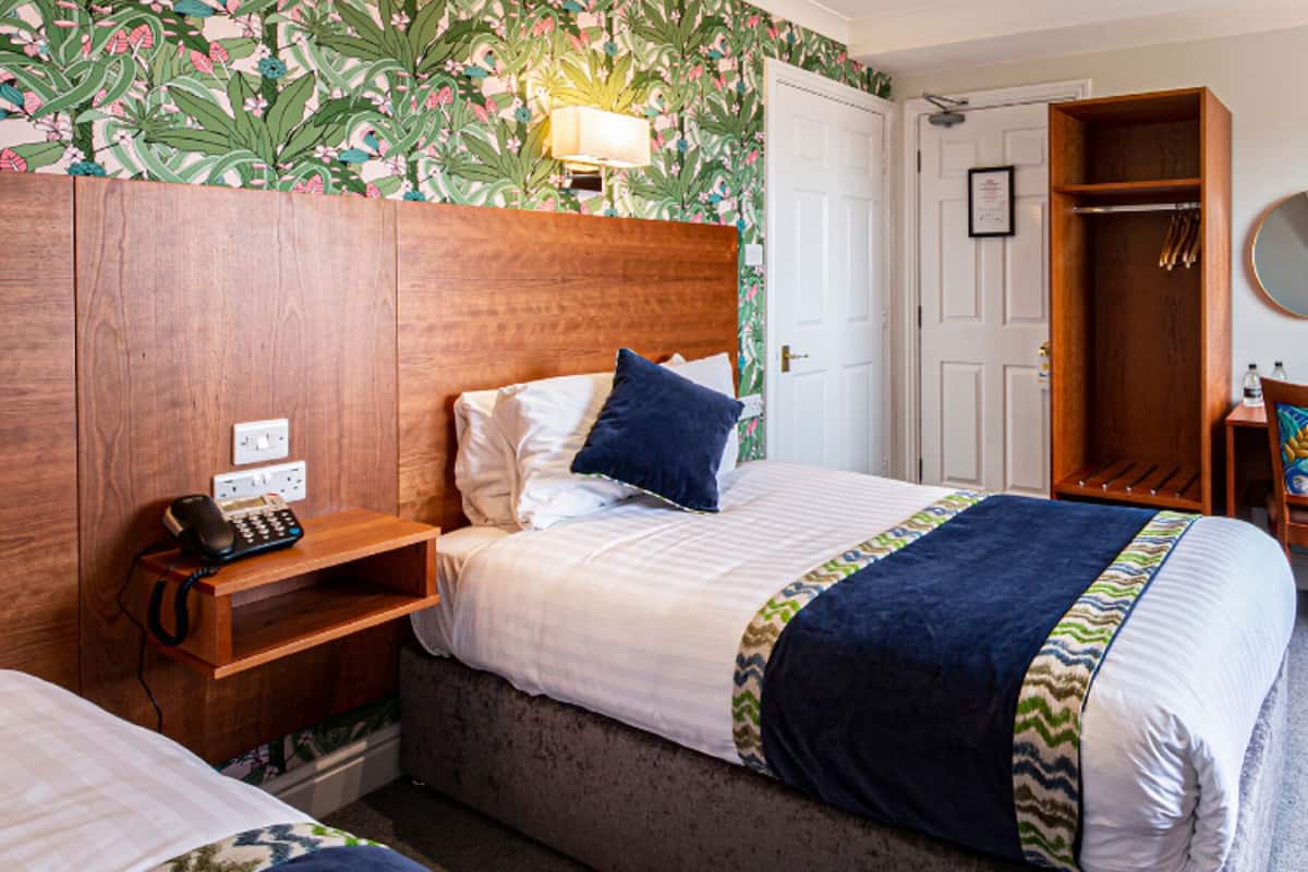 A hotel room with a bed, bedside table, chair, and a small desk with a mirror. There is a door leading to a bathroom and a window providing natural light.