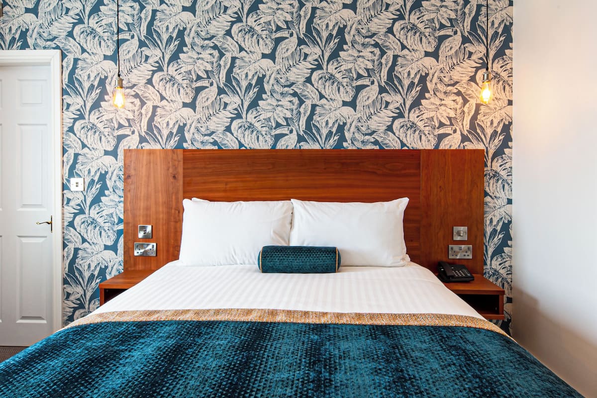 A hotel room with a wooden headboard, white bedding, a teal throw blanket, and a floral wallpaper pattern.