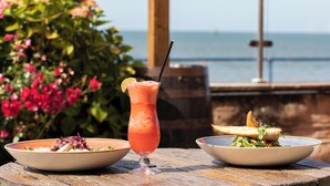 Une table dressée avec une boisson, une salade et du pain, le tout placé devant une table en bois et avec une vue sur la mer.
