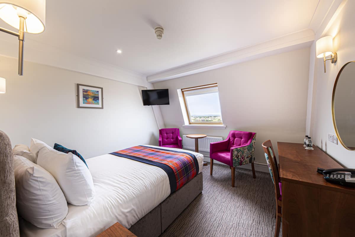 A hotel room with a bed, a desk with a telephone, a chair, a small table, a television mounted on the wall, and a window with a view.