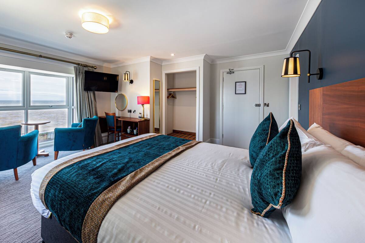 A hotel room with a large bed, a desk with a chair, a TV mounted on the wall, and a view of the ocean.