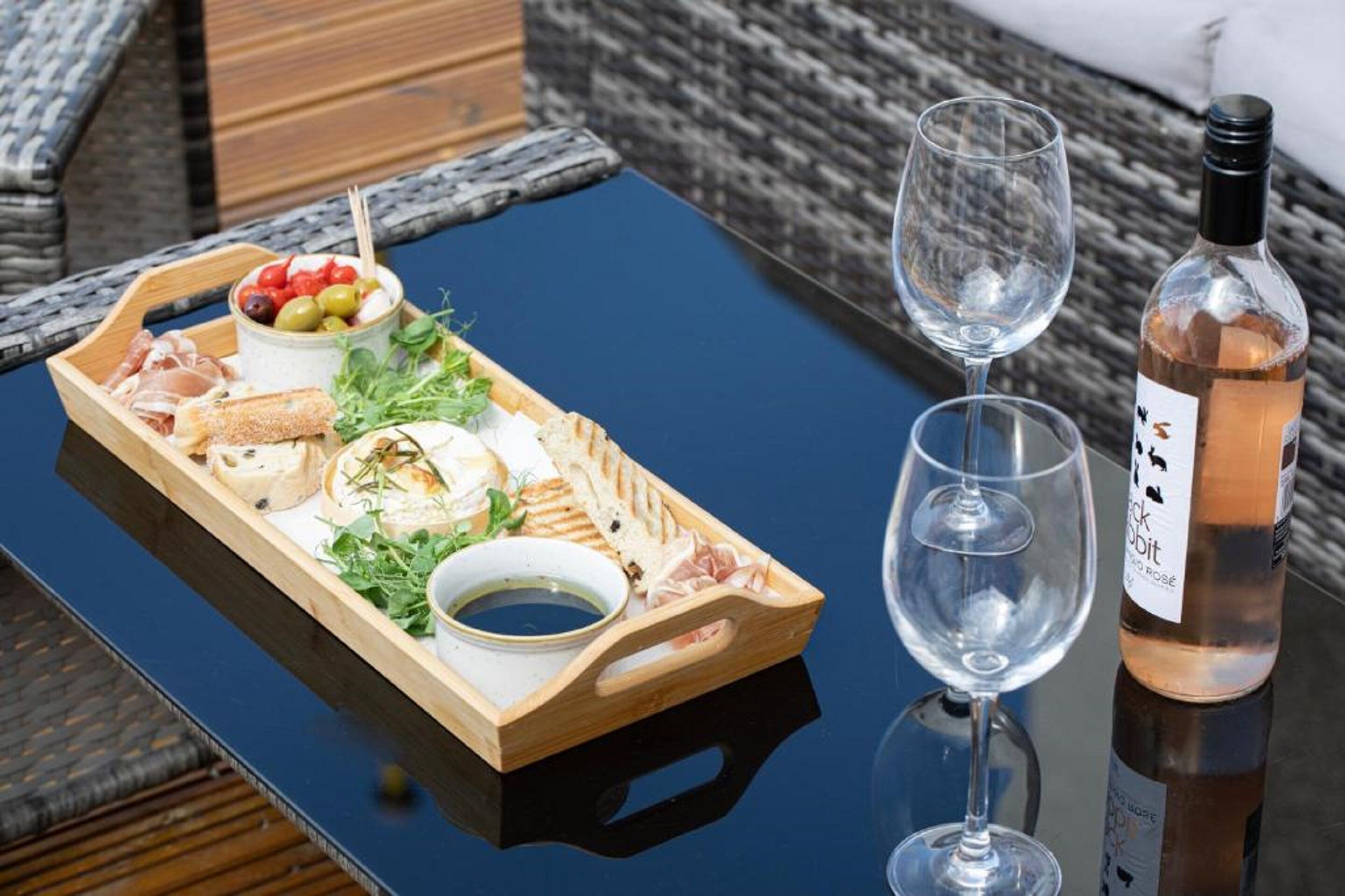 A wooden tray with various cheeses, olives, and greens on a table, accompanied by two wine glasses and a bottle of wine.