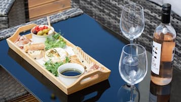 A wooden tray with various cheeses, olives, and greens on a table, accompanied by two wine glasses and a bottle of wine.