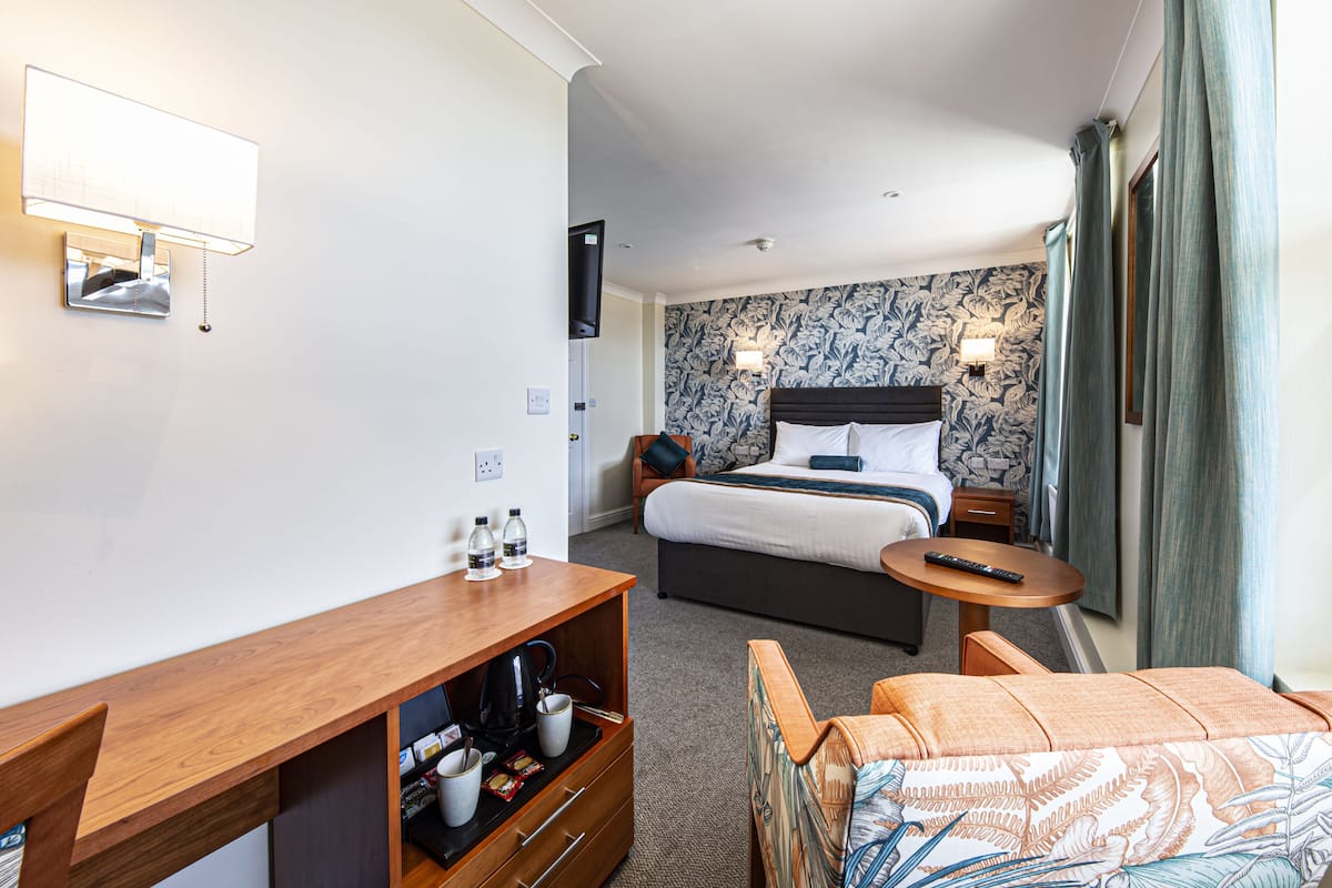 A hotel room with a bed, a desk with a coffee maker, a TV, and a wall-mounted lamp.