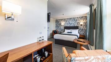 Quarto de hotel com cama, escrivaninha com cafeteira, televisão e luminária na parede.