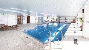 Une grande piscine intérieure à l’eau d’un bleu limpide, entourée de chaises longues et de plantes en pot.