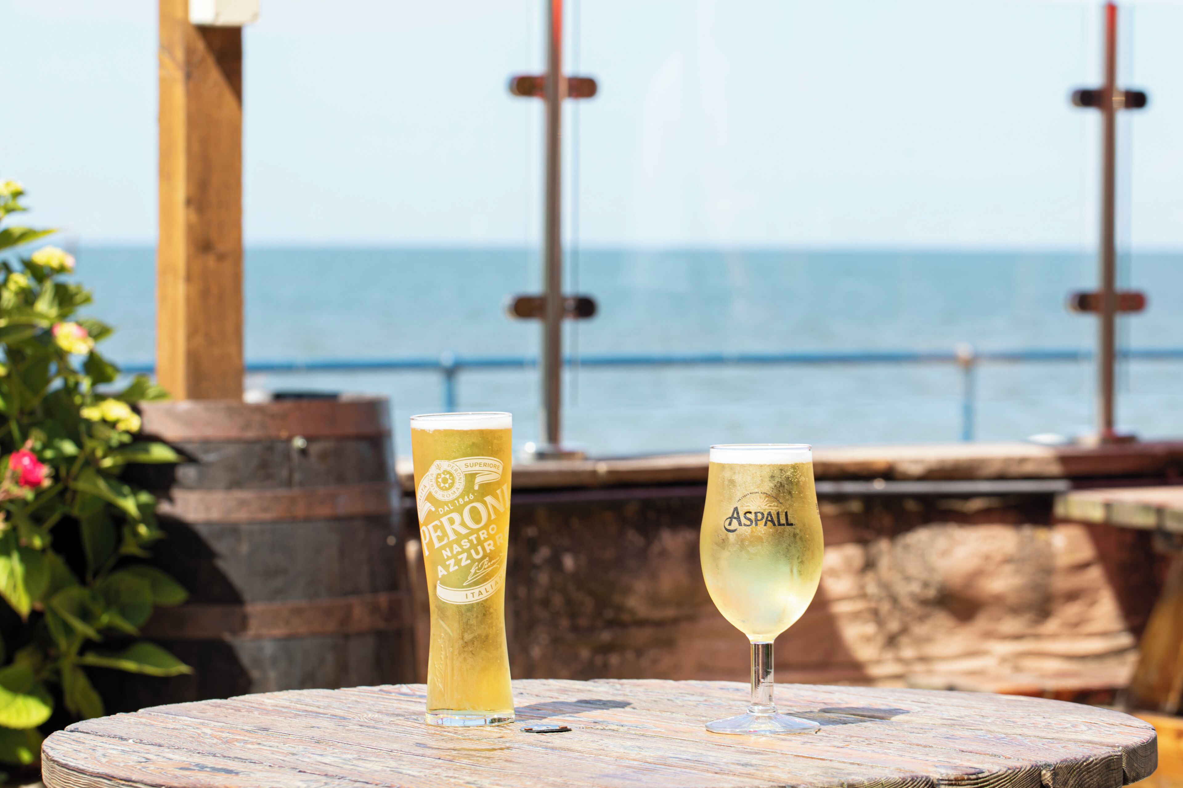 Two glasses of beer on a wooden table with a view of the sea in the background.