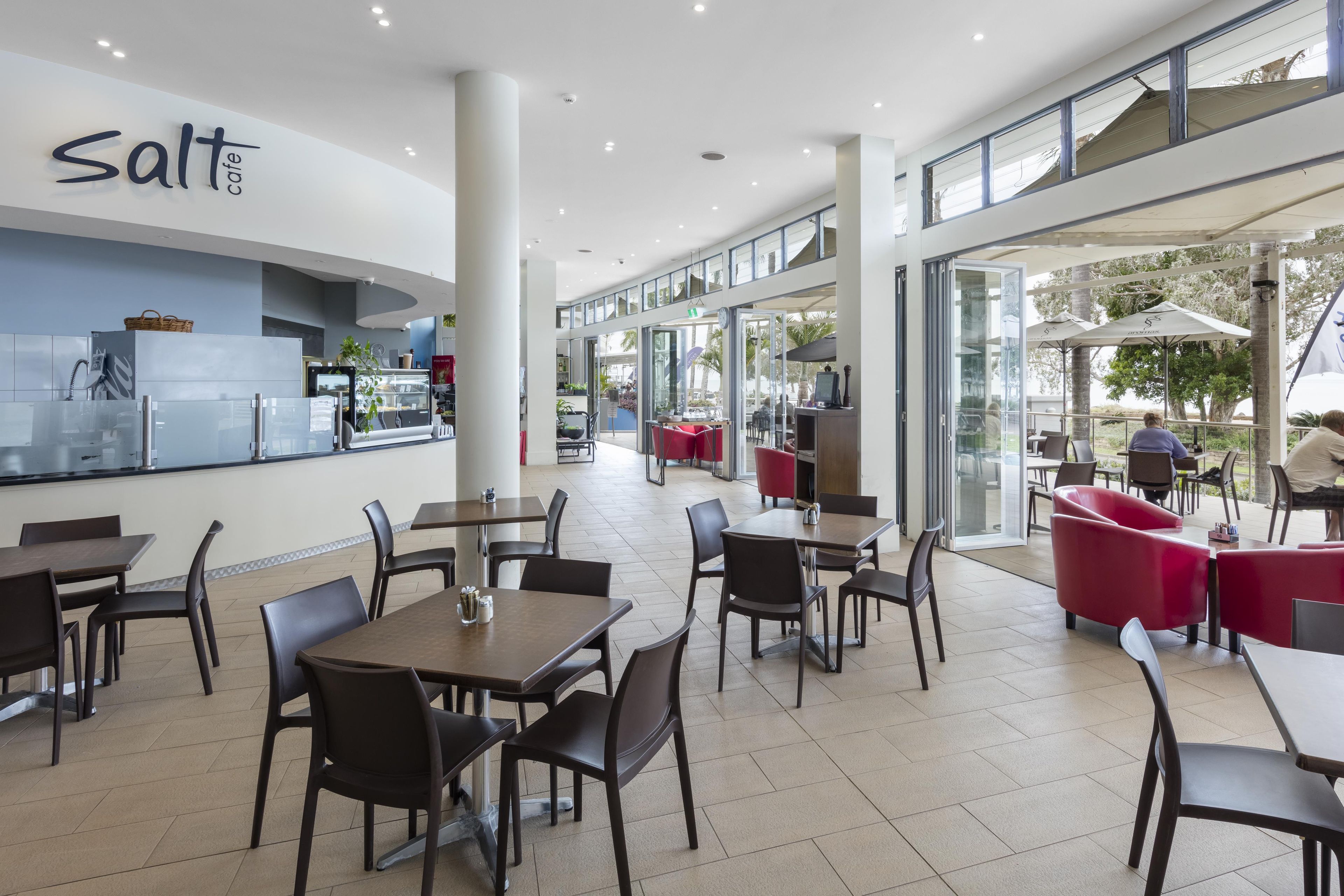 Interior moderno de una cafetería con mesas y sillas, una barra con dispensador de bebidas y vistas al exterior.
