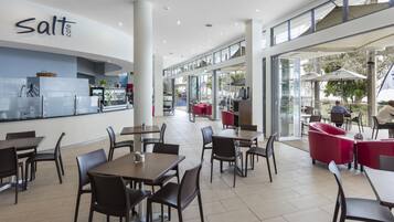Interior moderno de una cafetería con mesas y sillas, una barra con dispensador de bebidas y vistas al exterior.