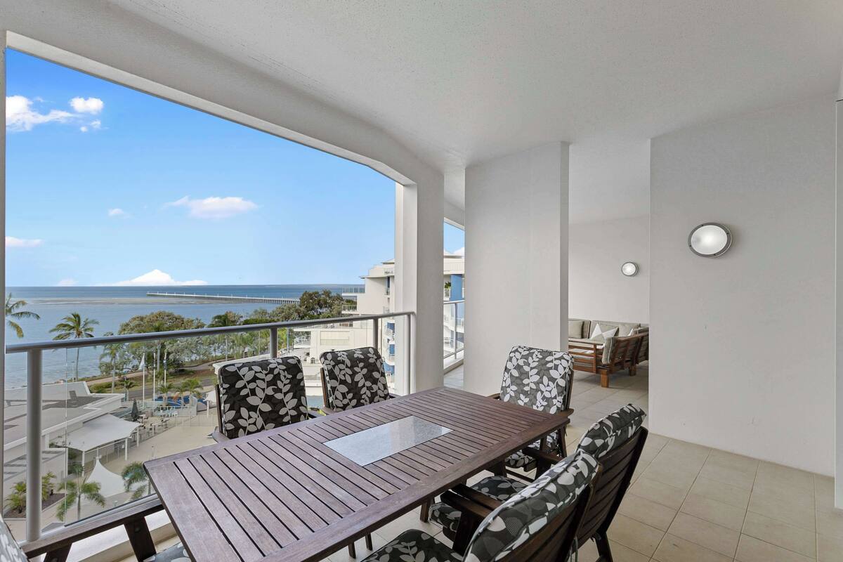 Un balcón con mesa y sillas de madera, con vista a una playa y al océano.