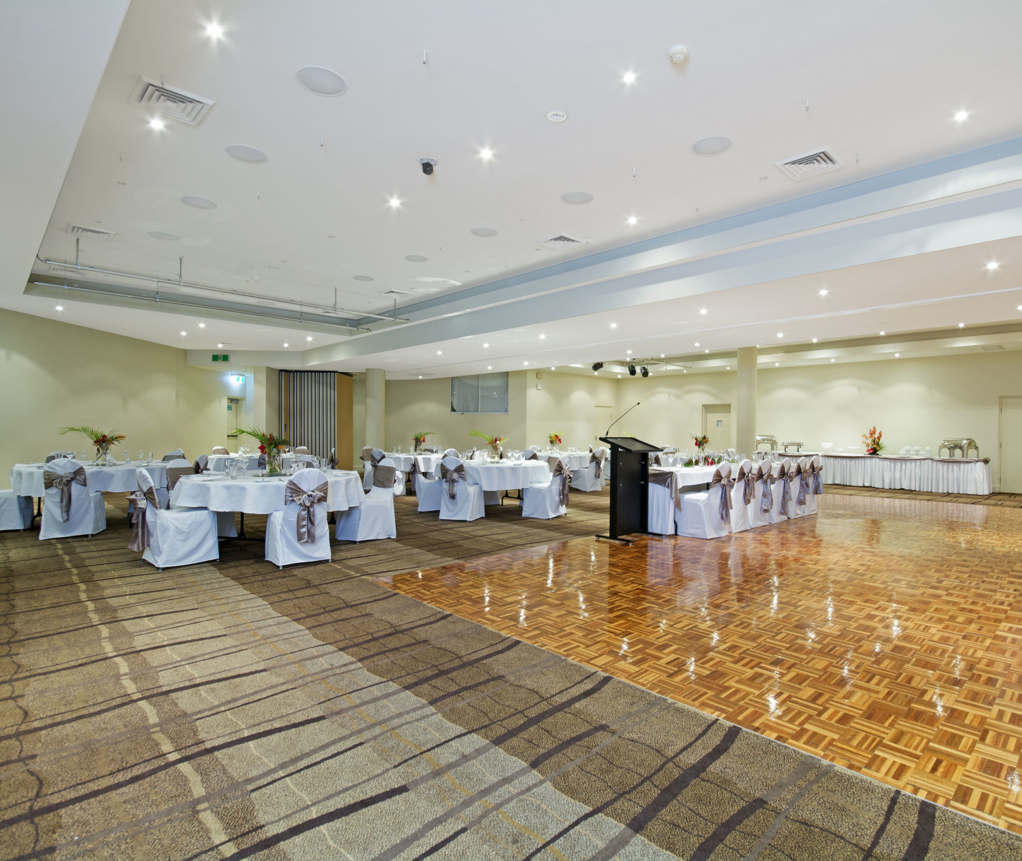 Une grande salle de banquet, bien éclairée, avec de nombreuses tables rondes recouvertes de nappes blanches et des chaises ornées de rubans gris.