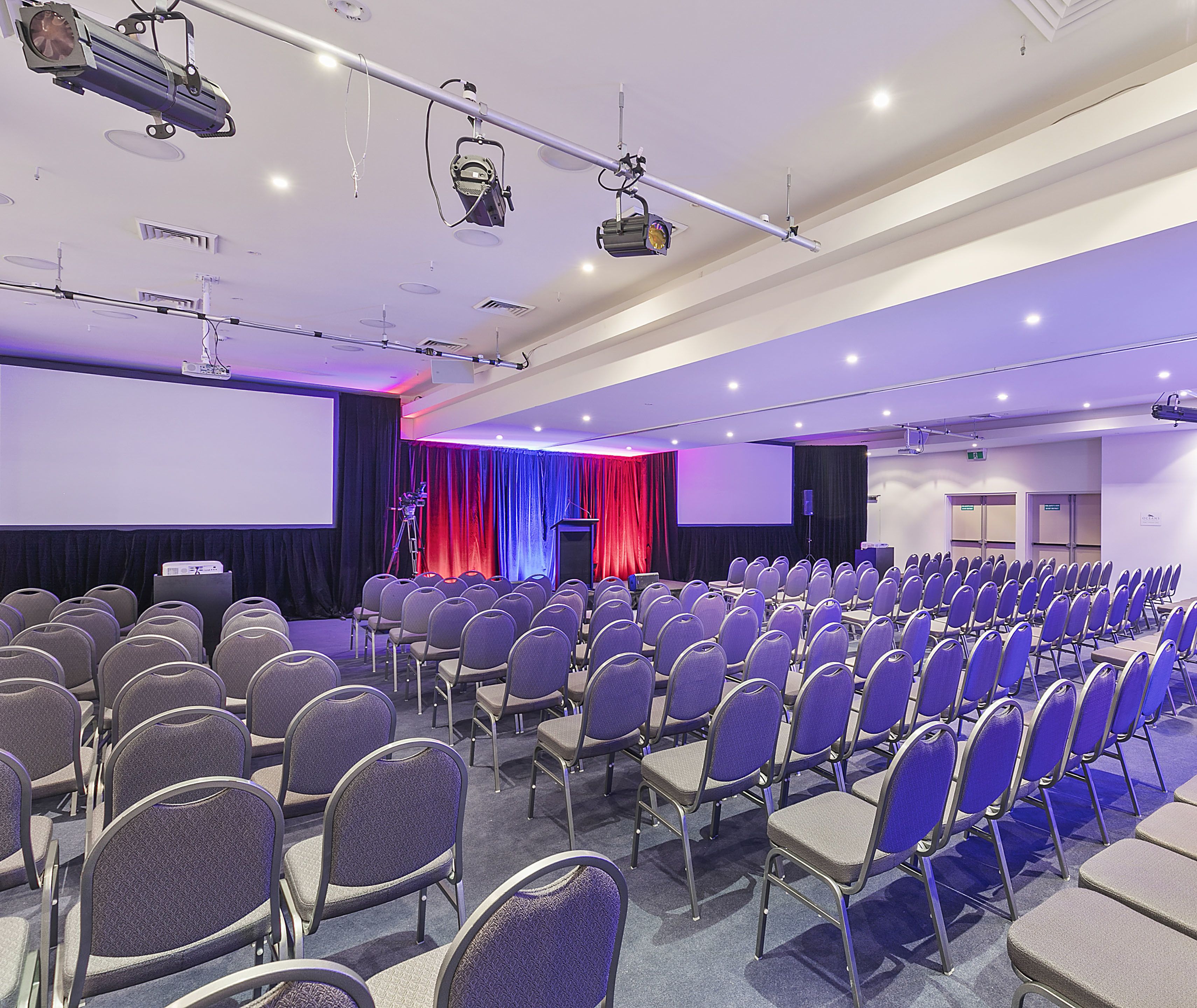 A conference room with rows of chairs, a stage with a podium, and a large screen.