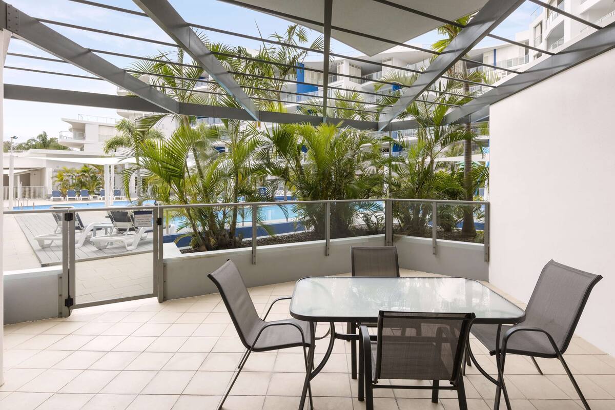 Un patio exterior moderno con una mesa y sillas de vidrio, rodeado de barandas de metal y palmeras.