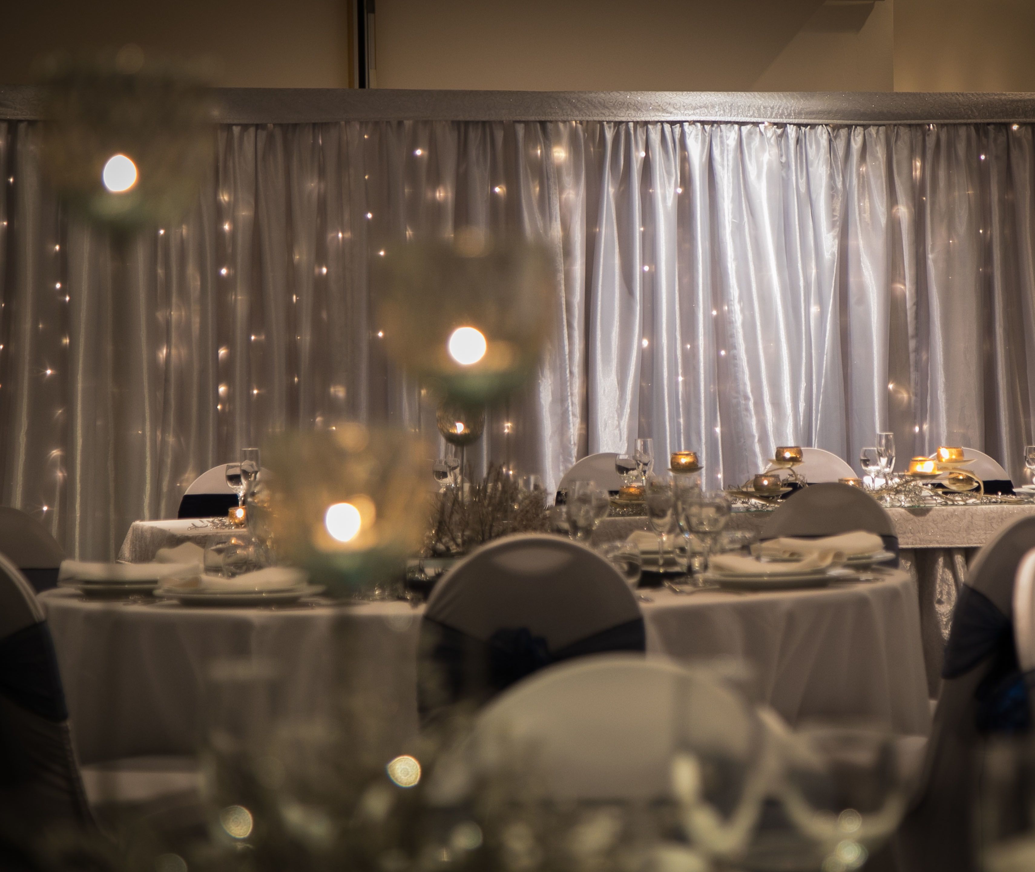 A formal dining room with tables set for a meal, featuring a backdrop of sheer curtains and soft lighting.