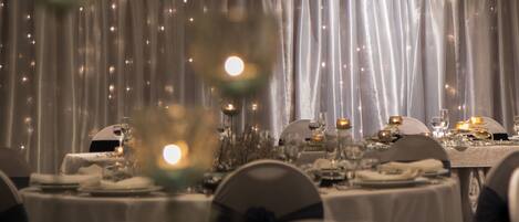 A formal dining room with tables set for a meal, featuring a backdrop of sheer curtains and soft lighting.