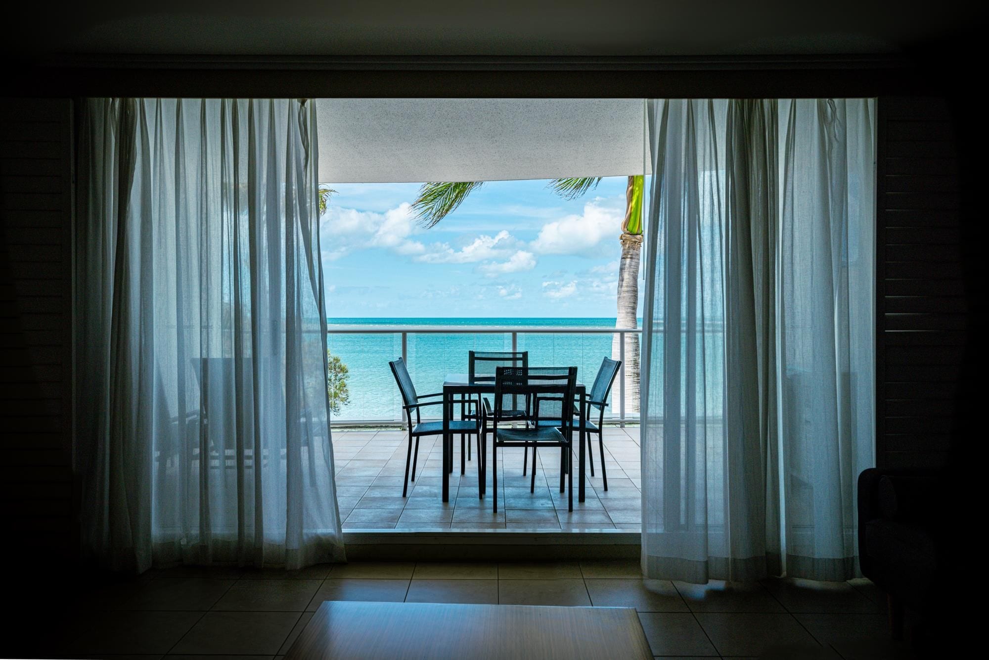 A room with a view of the ocean through sheer curtains, a small table with chairs, and a tiled floor.