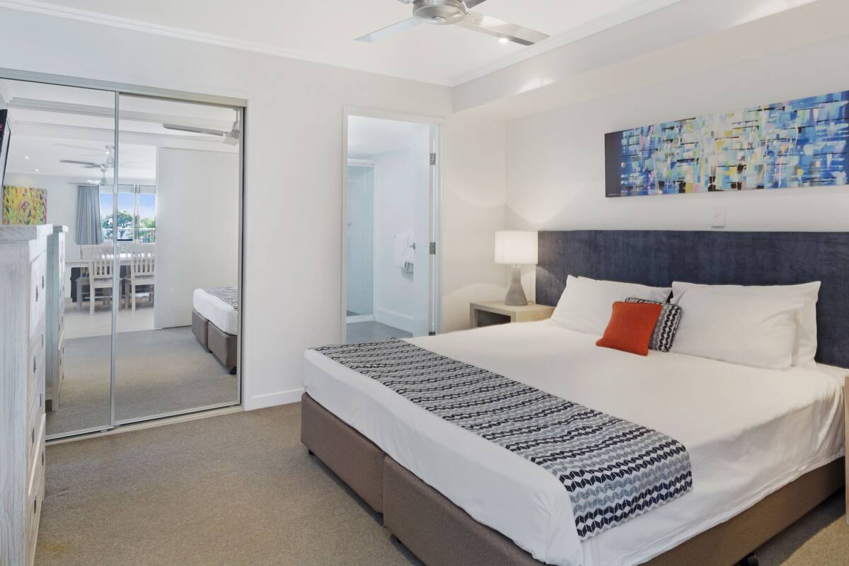 Une chambre d’hôtel moderne avec un grand lit, un ventilateur de plafond, une porte vitrée donnant sur un balcon et un tableau au mur.