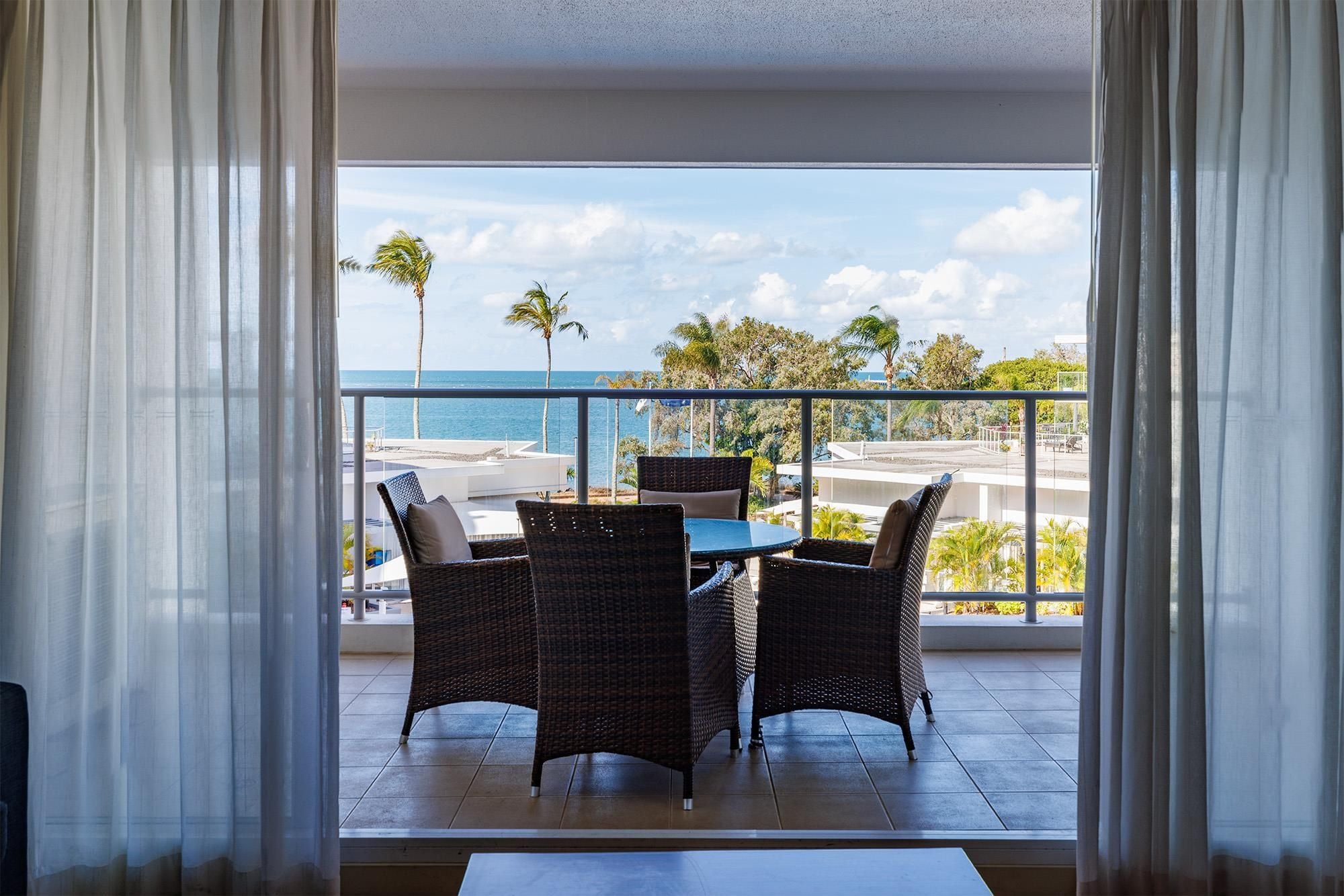 A balcony with wicker chairs and a table, overlooking a beach and ocean.