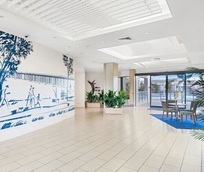 Een ruime lobby met een betegelde vloer, een groot muurschildering dat een pierafbeelding toont, en een zithoek met tafels en stoelen.