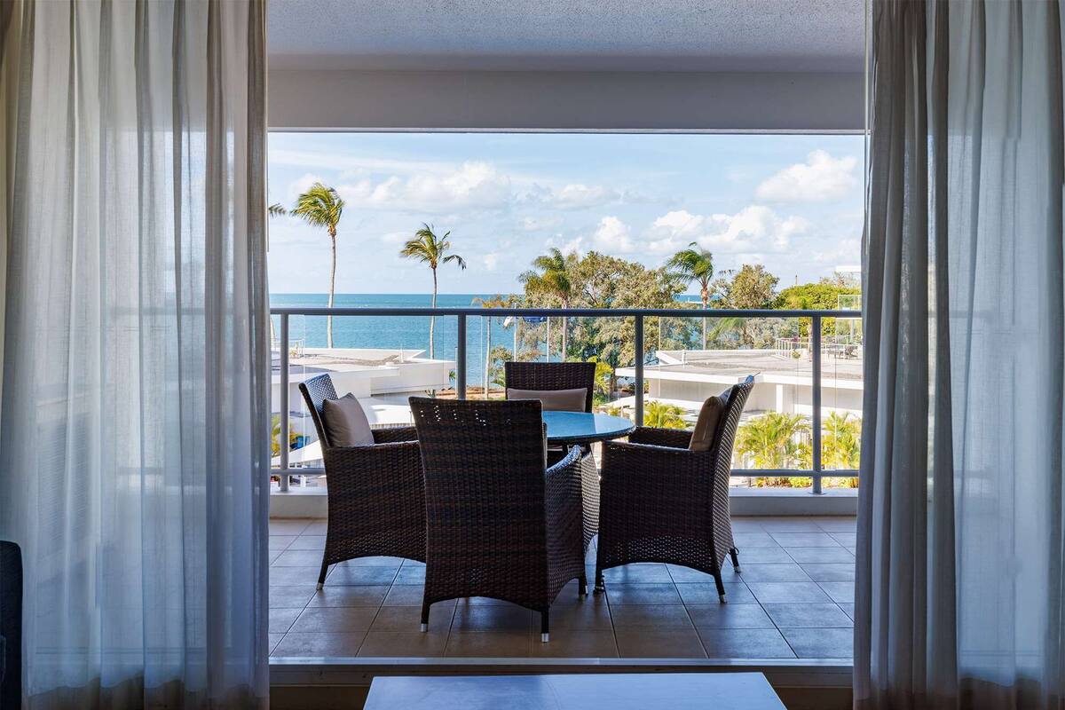 Un balcón con sillones de mimbre y una mesa, con vista a una playa y palmeras.