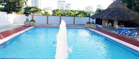Una piscina con fuente de agua, sillas de descanso y una estructura de techo de paja.