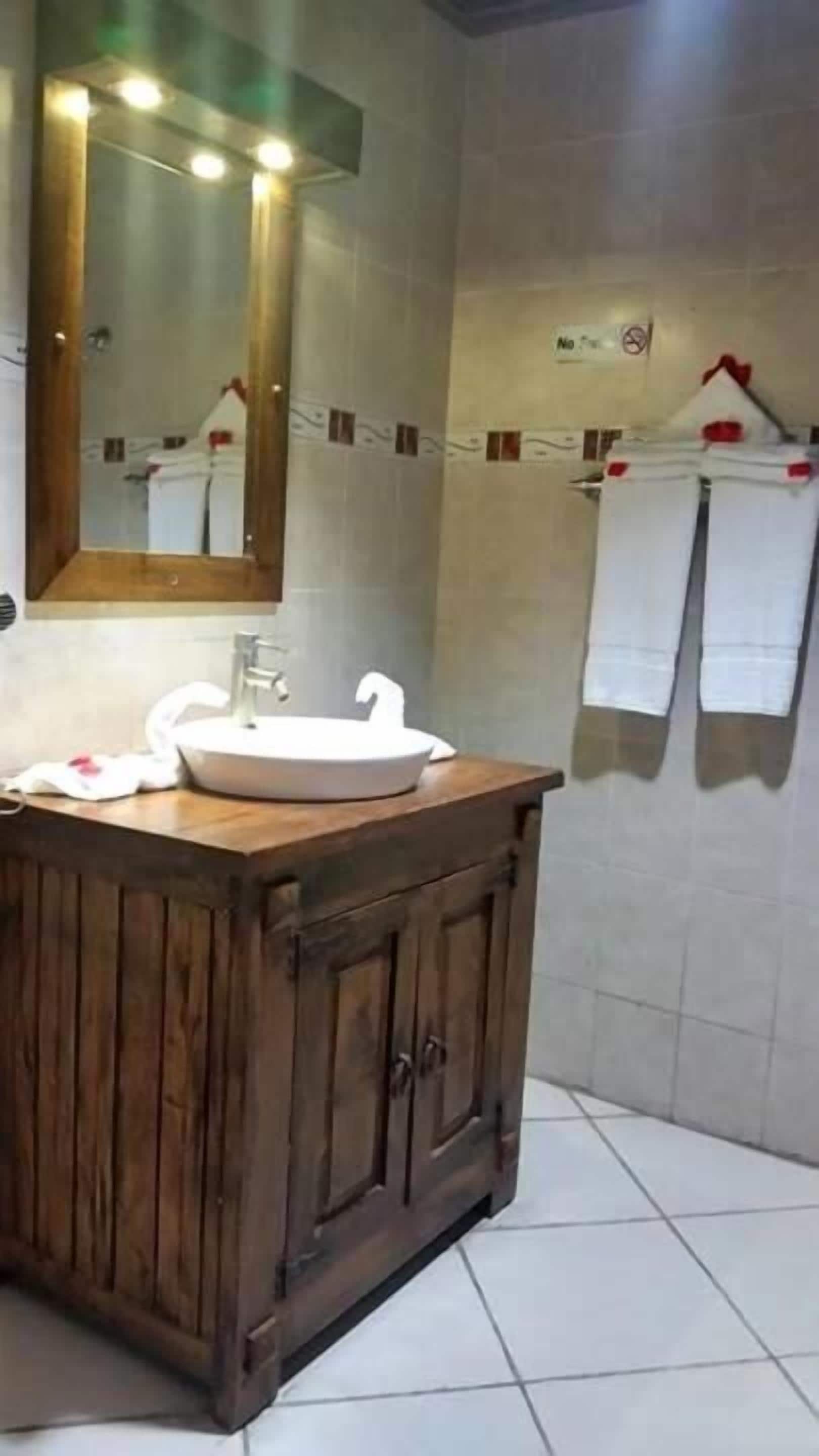 A bathroom with a wooden vanity, a white sink, and a mirror above it. There are towels hanging on a rack and a no-smoking sign on the wall.