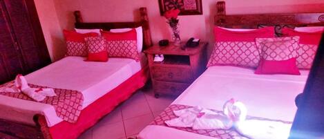 Two double beds with red and white bedding, a wooden nightstand with a telephone, and a vase with red flowers.