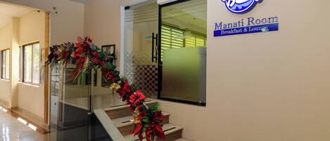 Un escalier doté d’une rampe en verre et décoré de motifs rouges et verts, menant à une salle dont la porte indique « Salle Manati – Petit-déjeuner et salon ».