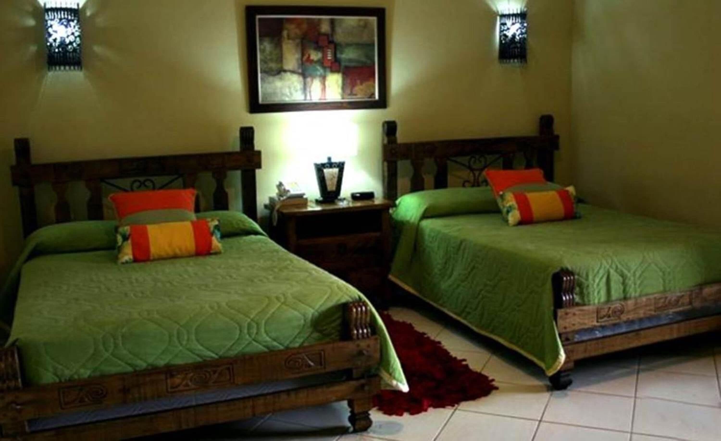 Two single beds with wooden frames and green bedding, a nightstand with a lamp, and a painting on the wall.