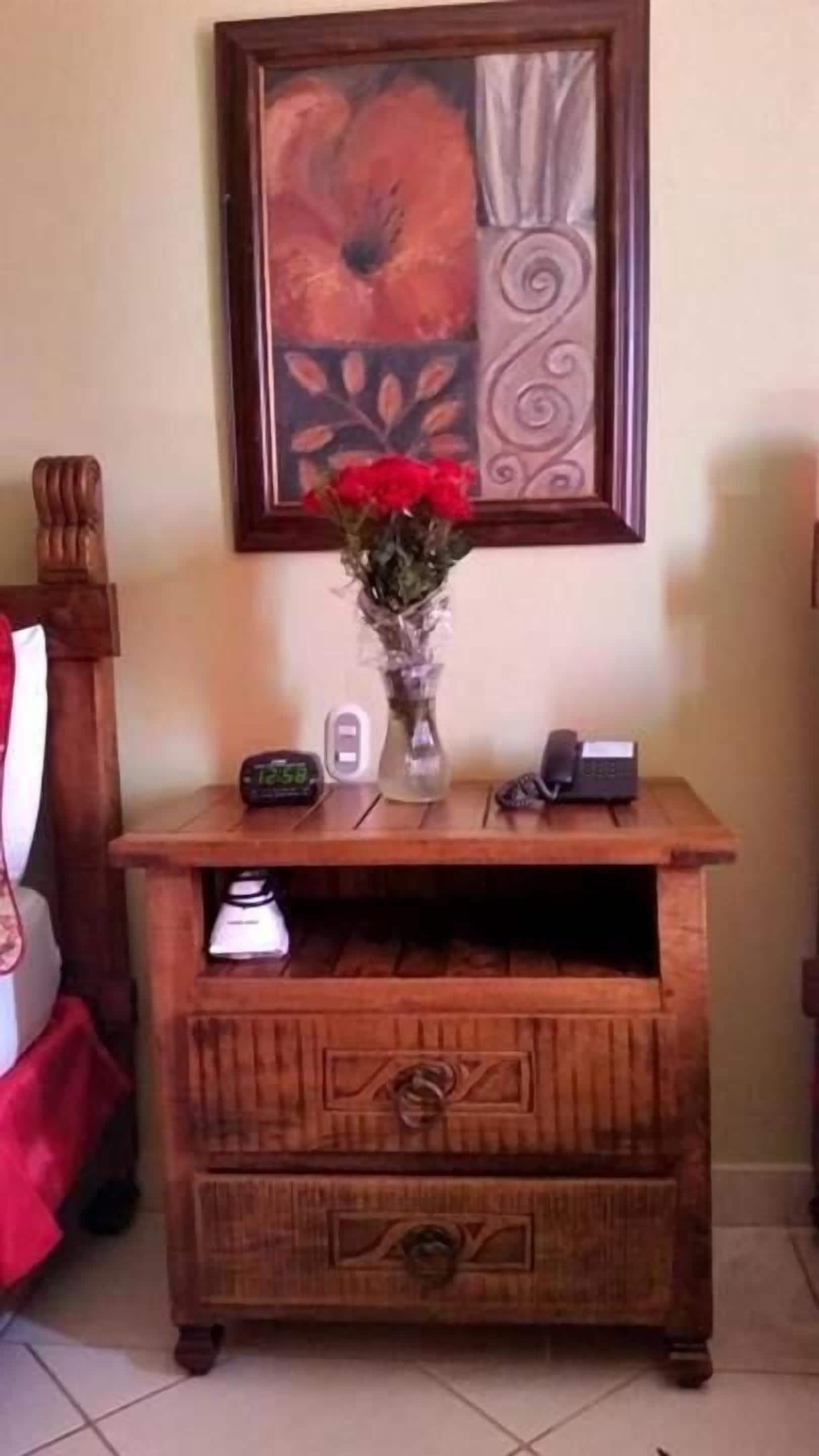 Un mueble de madera con un jarrón de flores rojas, un reloj y un pequeño dispositivo electrónico encima.