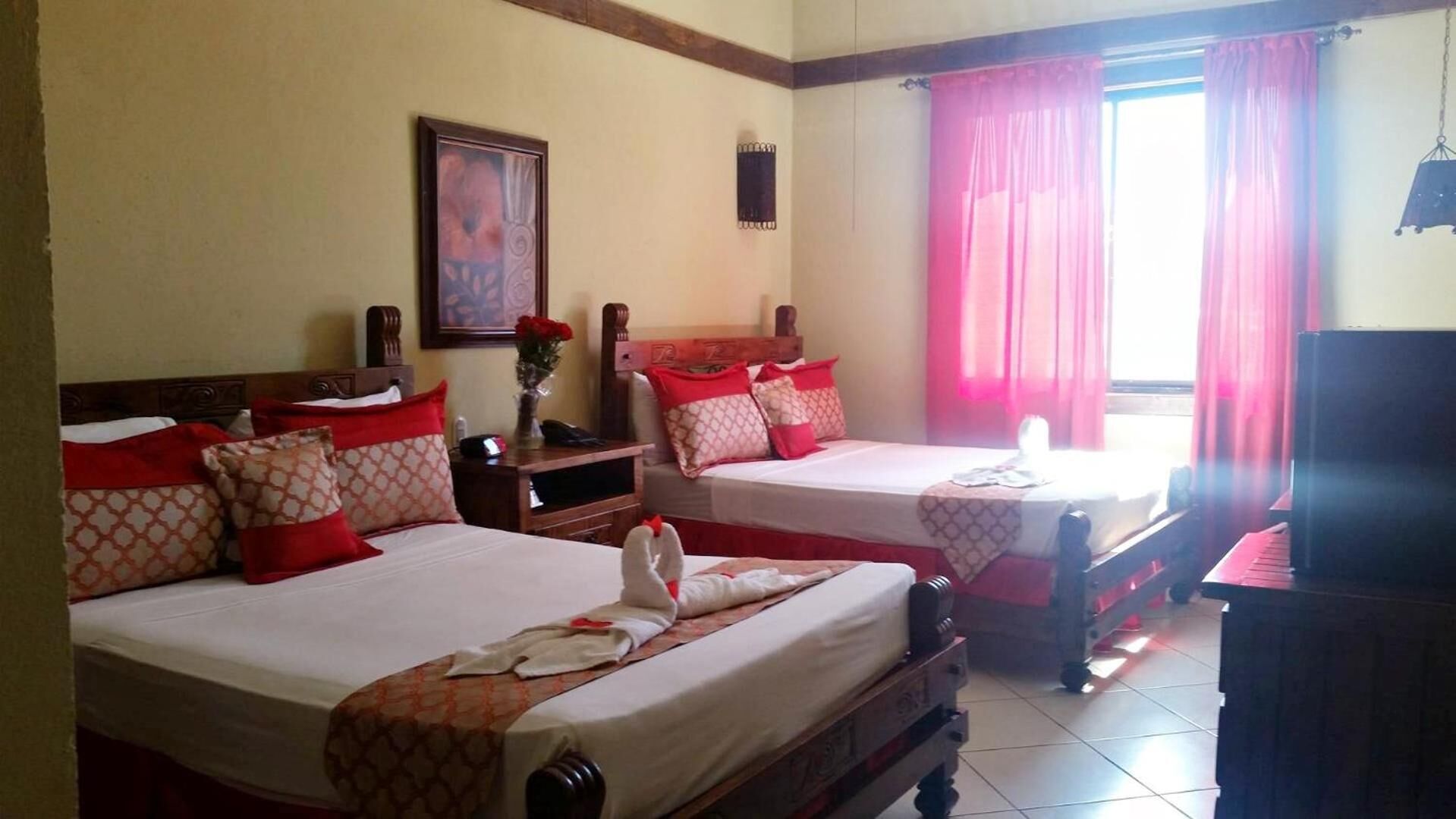 A hotel room with two beds, each with red and patterned pillows, a wooden headboard, a nightstand, a framed picture on the wall, and a window with pink curtains.