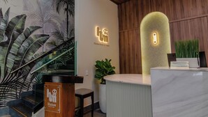 Un área de recepción con un mostrador de madera, una planta grande en maceta y una pared con un mural tropical.