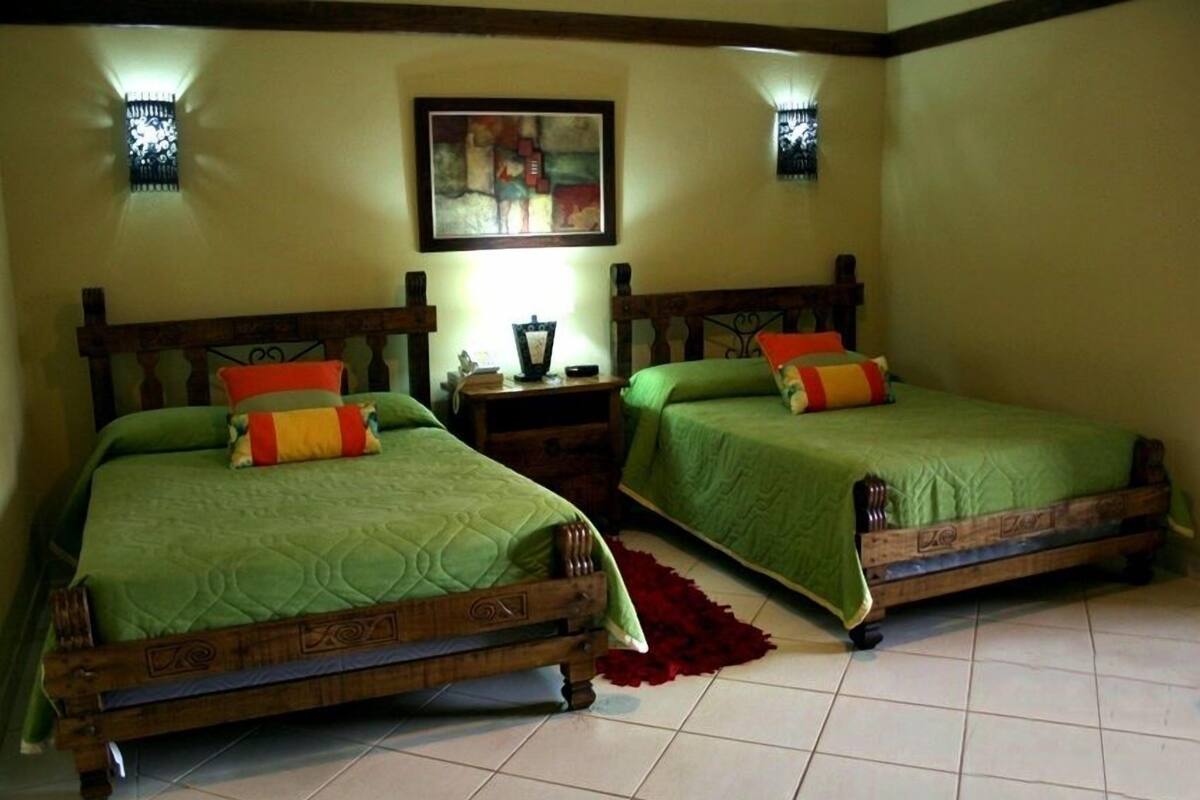 Two wooden beds with green bedding and red pillows, a nightstand with a lamp, and a framed abstract painting on the wall.