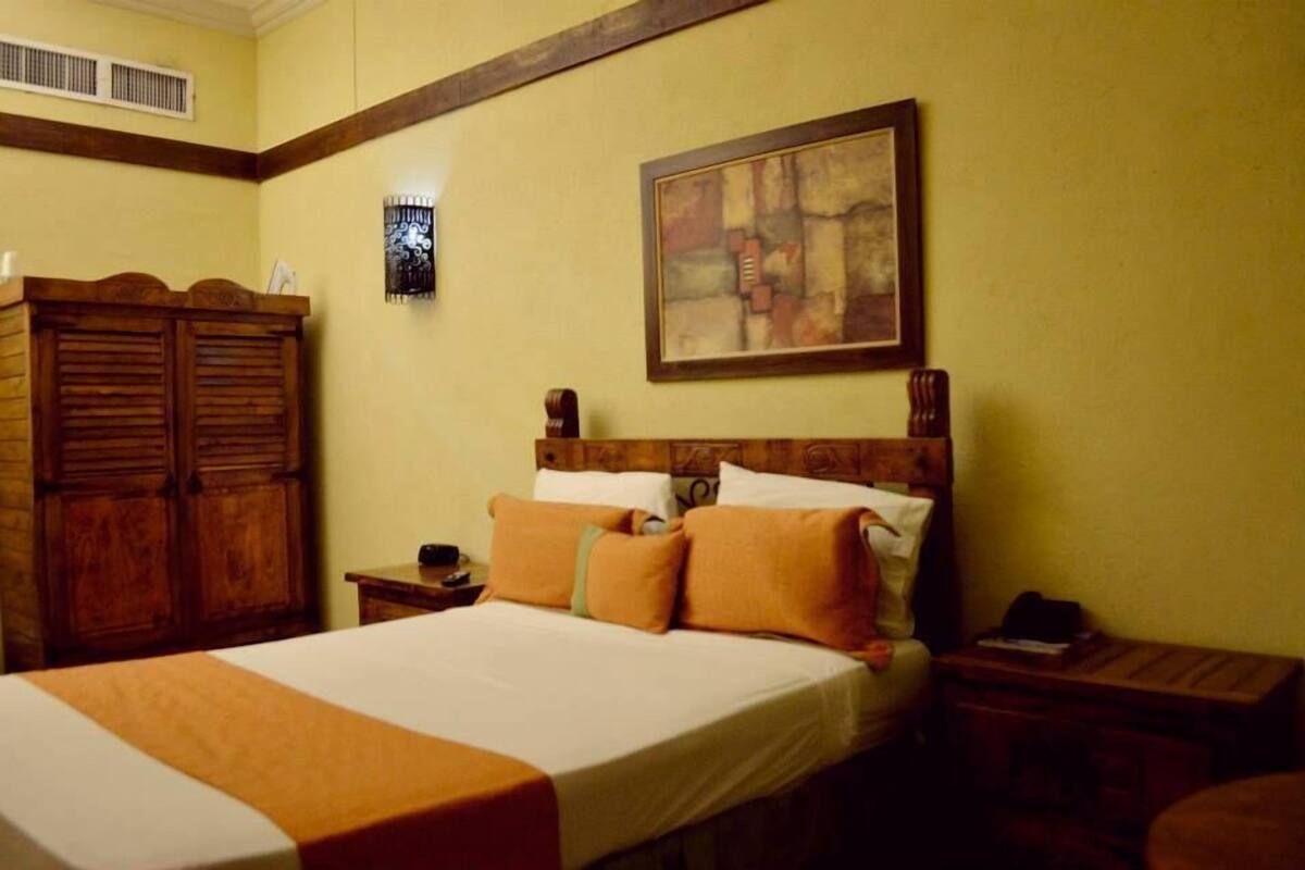 A hotel room with a large bed, wooden wardrobe, a painting on the wall, and a small table with a lamp.