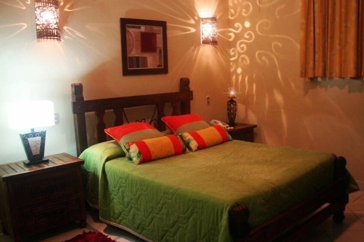 A bedroom with a green bedspread, two pillows, a wooden headboard, a nightstand with a lamp, and wall sconces.