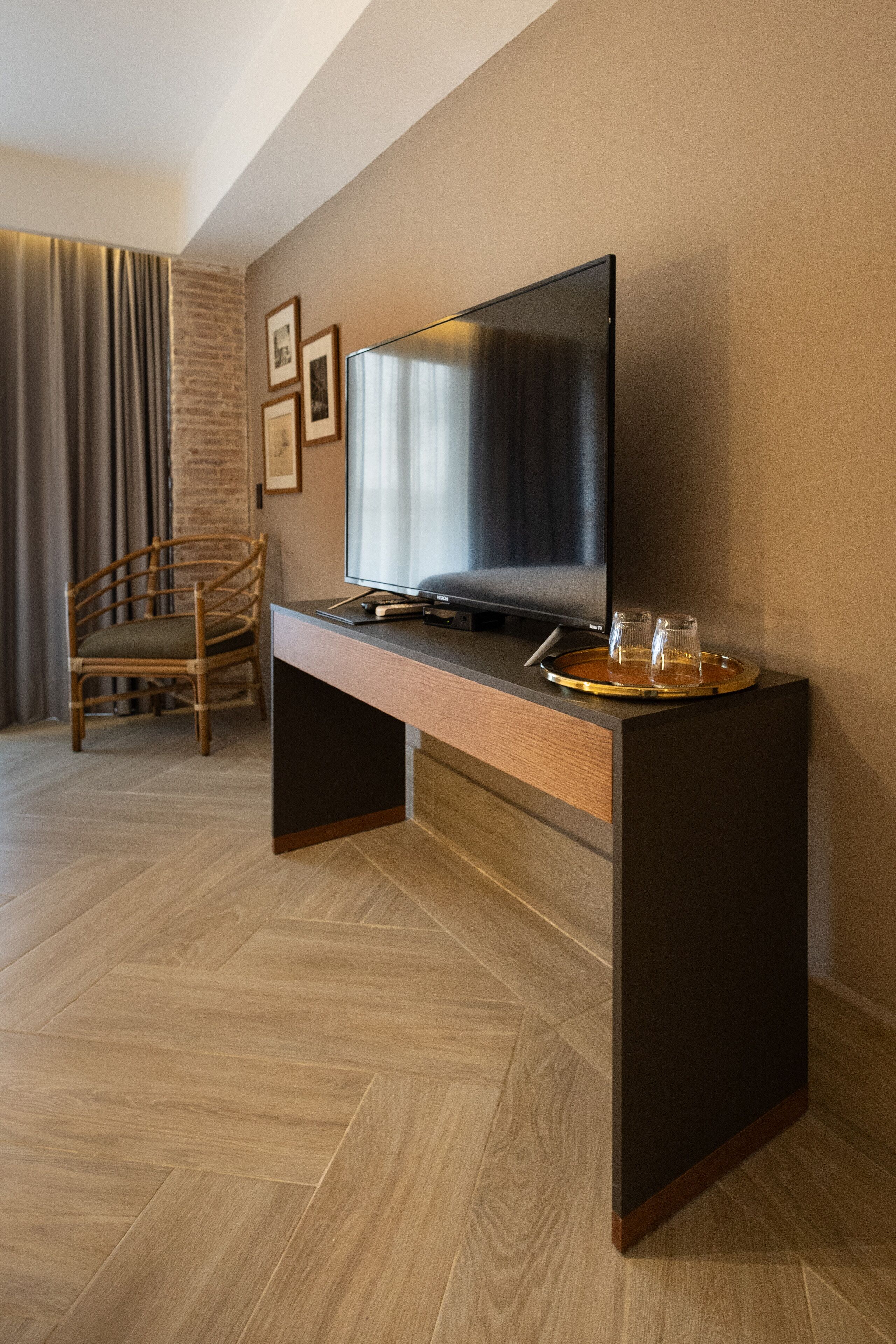 Quarto moderno com piso de madeira em padrão herringbone, uma escrivaninha escura com televisão de tela plana, uma cadeira de vime e um tabuleiro com copos e um livro.