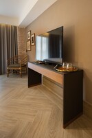 Quarto moderno com piso de madeira em padrão herringbone, uma escrivaninha escura com televisão de tela plana, uma cadeira de vime e um tabuleiro com copos e um livro.