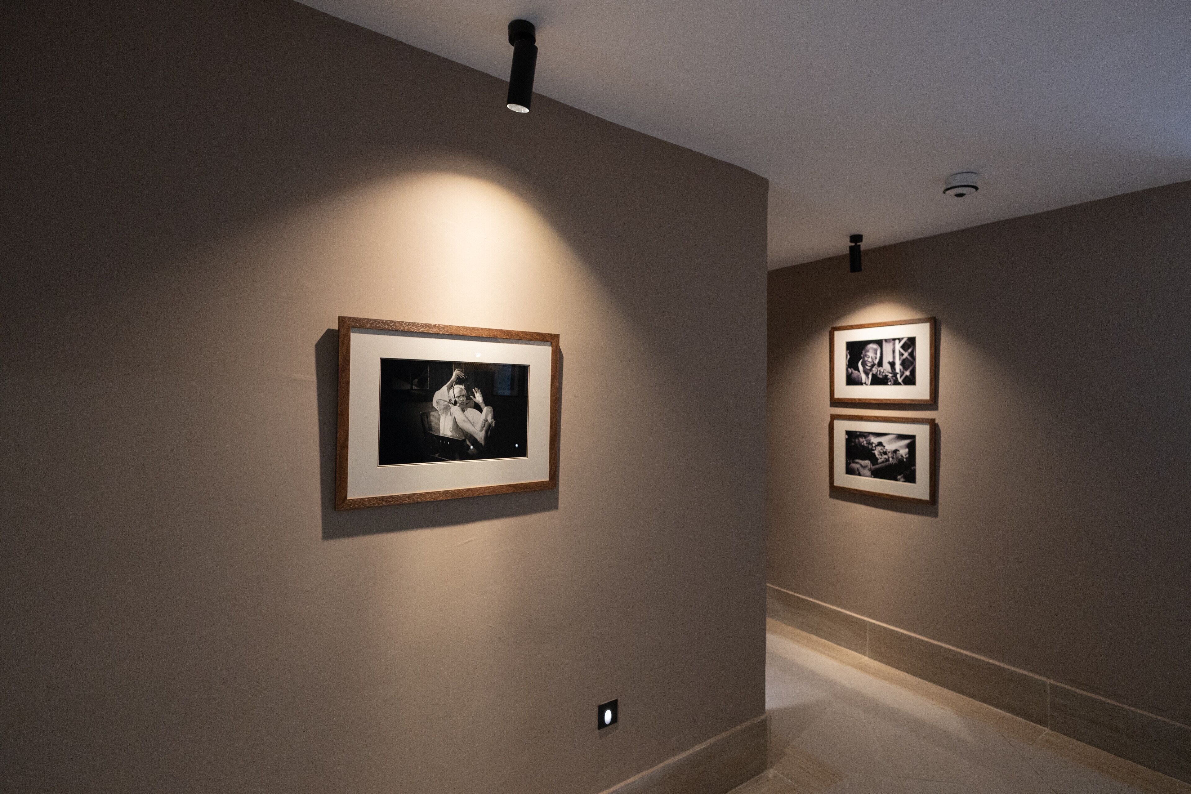 Een gang met ingelijste foto’s aan de muur, een lamp aan het plafond en een betegelde vloer.