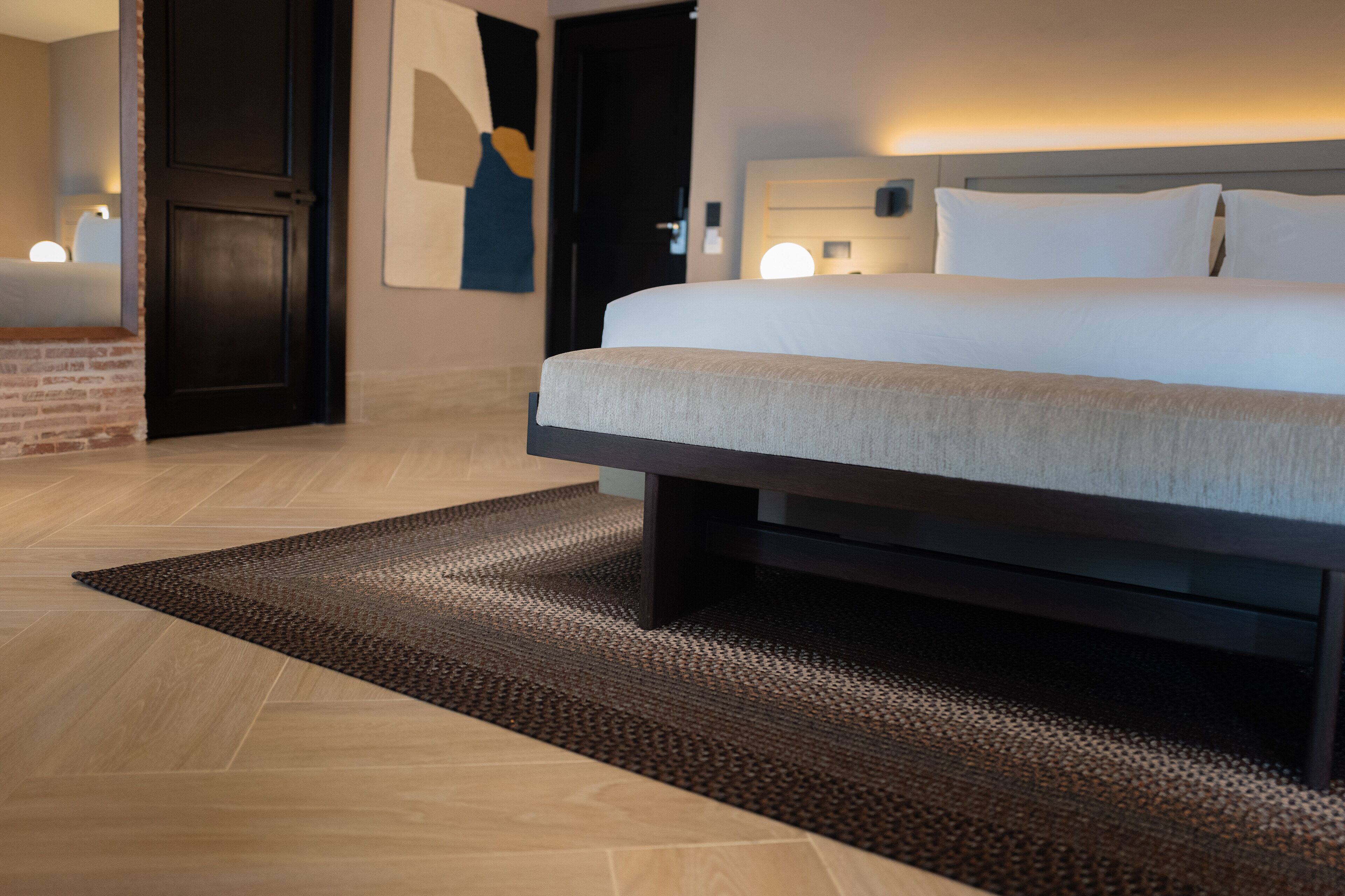 Quarto de hotel moderno com uma cama grande, uma cabeceira de madeira escura, um tapete texturizado e uma luminária fixada na parede.