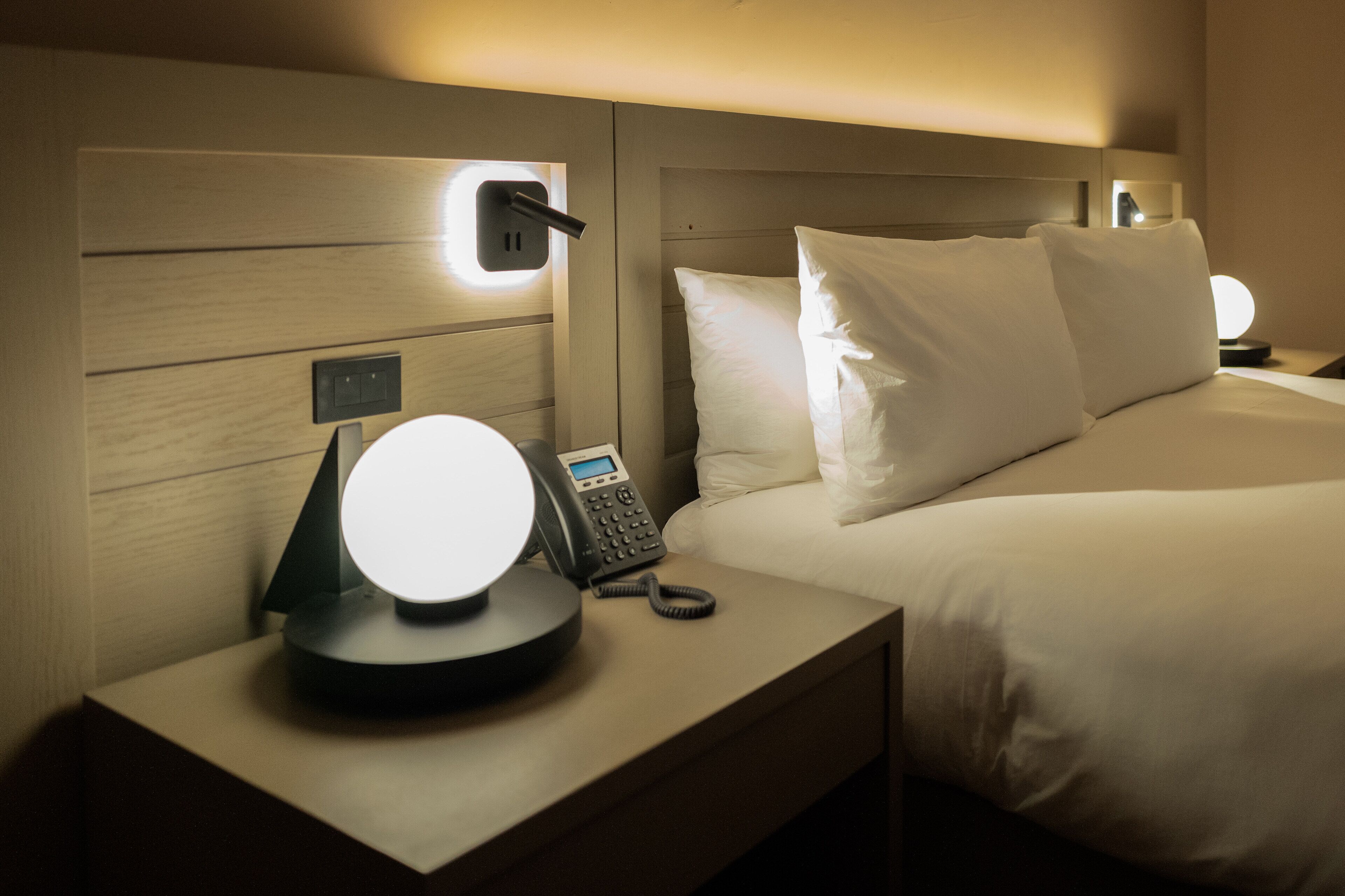Habitación de hotel con cabecera de madera, lámpara de noche, teléfono y cama con ropa blanca.
